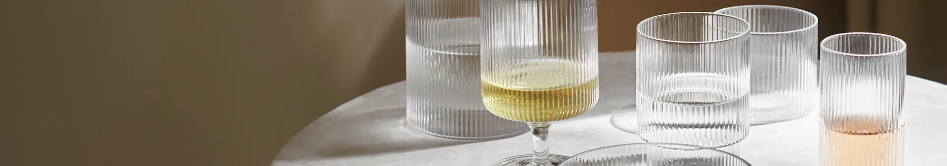 Rillet champagneglas med gul væske ved siden af tre riflede glas med klar og lyserød væske på et lyst bord.