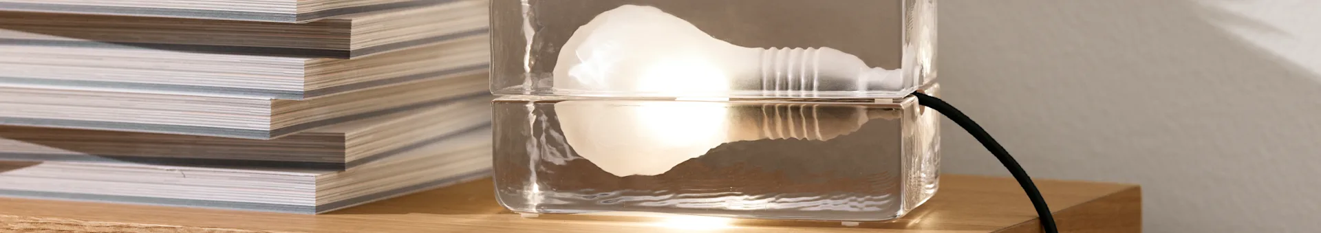 En unik, klar lampe, lavet af to stablede rektangulære blokke, med et glødende internt lys, ved siden af en stak magasiner på en træoverflade.