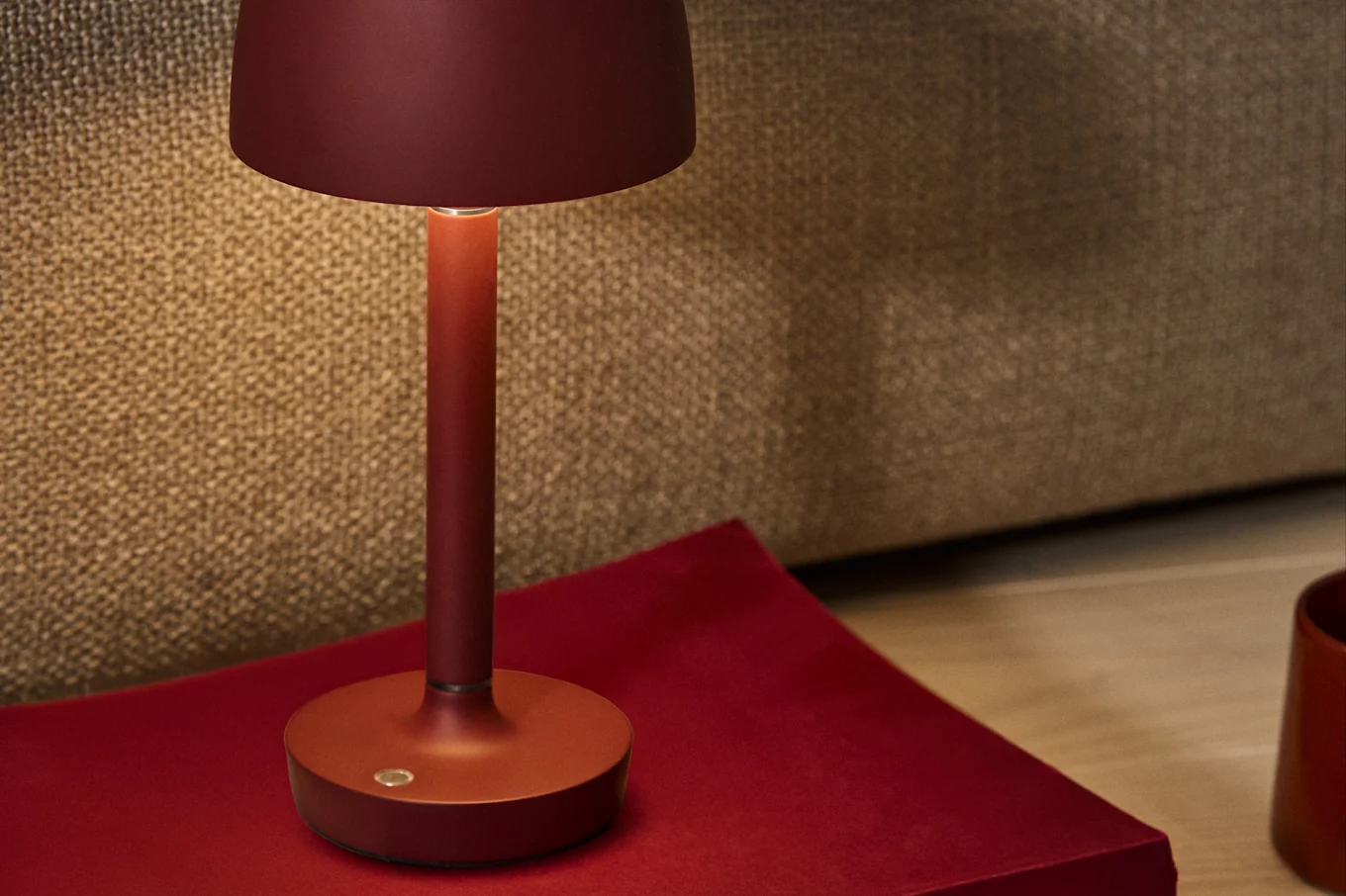 En dybrød minimalistisk bordlampe med et varmt skær, placeret på en rød overflade mod en tekstureret brun baggrund.