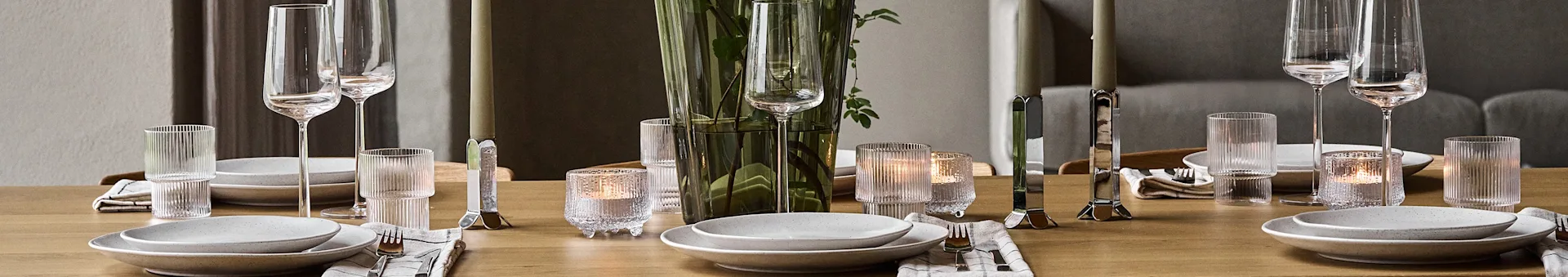Et træspisebord dækket med hvide tallerkener, sølvbestik, klare glas, tændte stearinlys og en grøn vase med blade.
