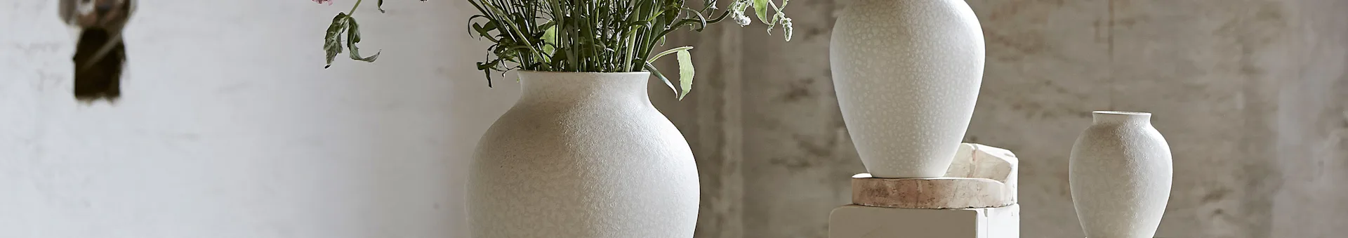 Tre teksturerede keramiske vaser i lyse farver, to med grønne planter og sarte blomster, mod en tekstureret væg.