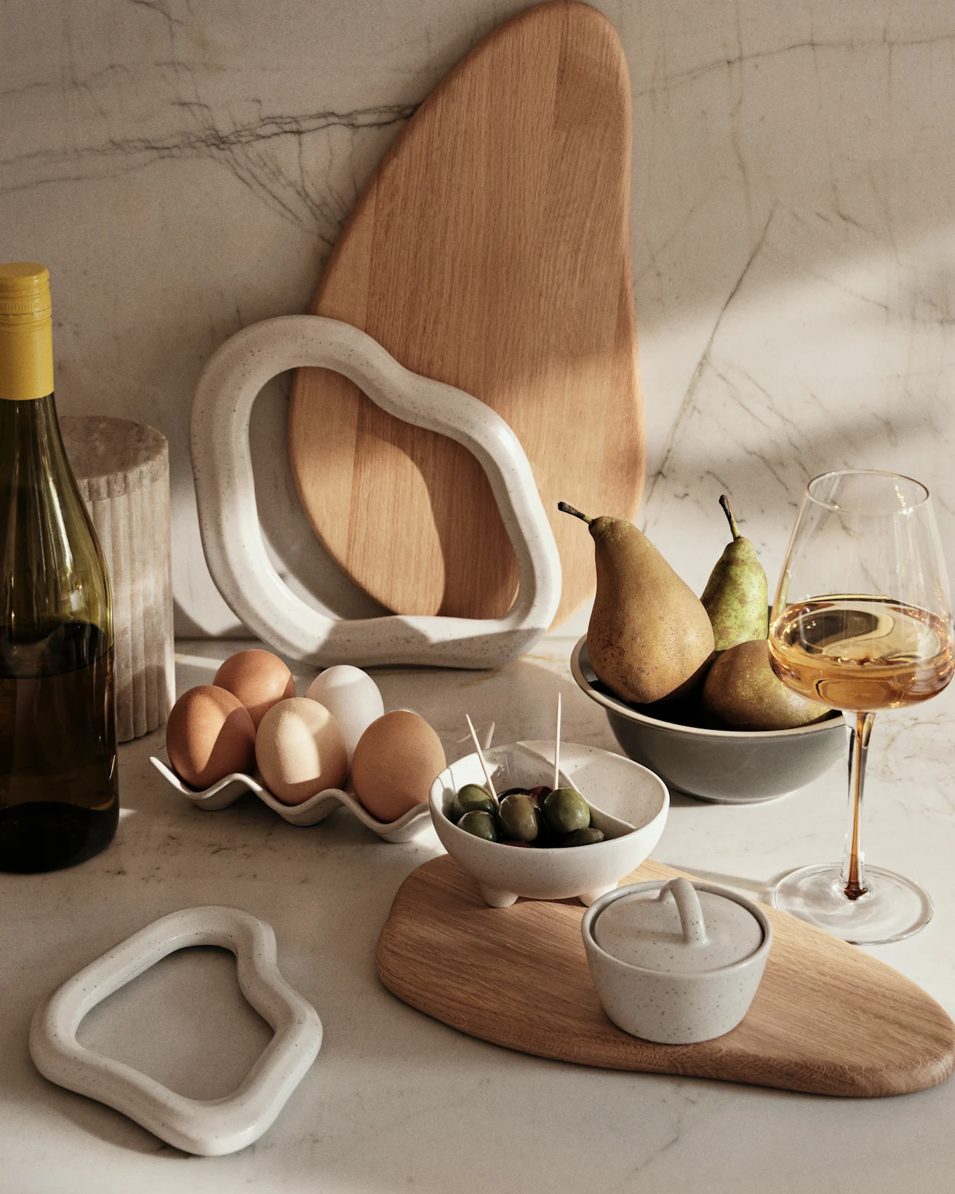 Marmorbord med vinflaske, æg, oliven, pærer og dekorative keramik- og trægenstande.