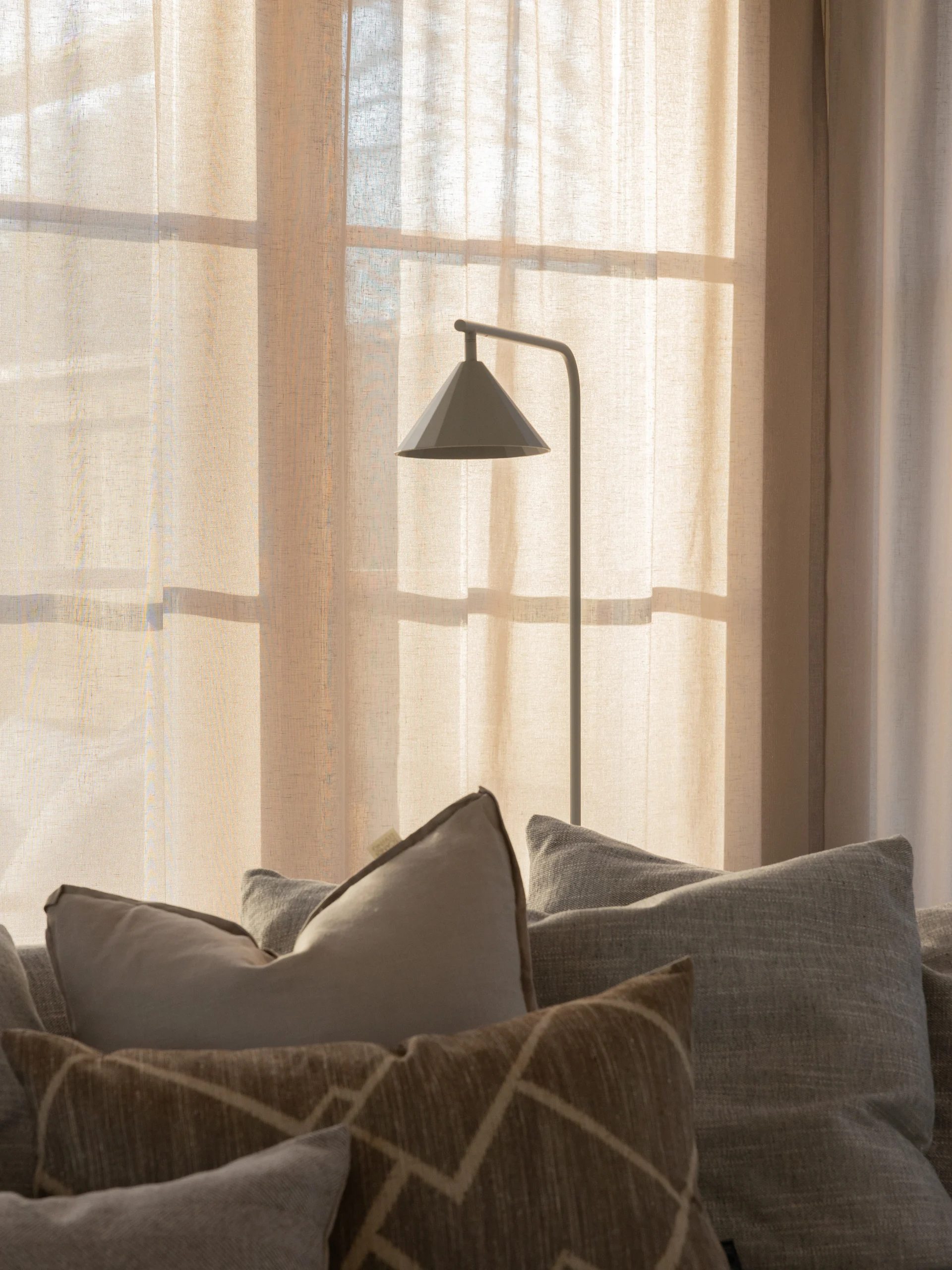 En moderne grå gulvlampe foran gennemsigtige, solbelyste gardiner, med forskellige puder på en sofa i forgrunden.
