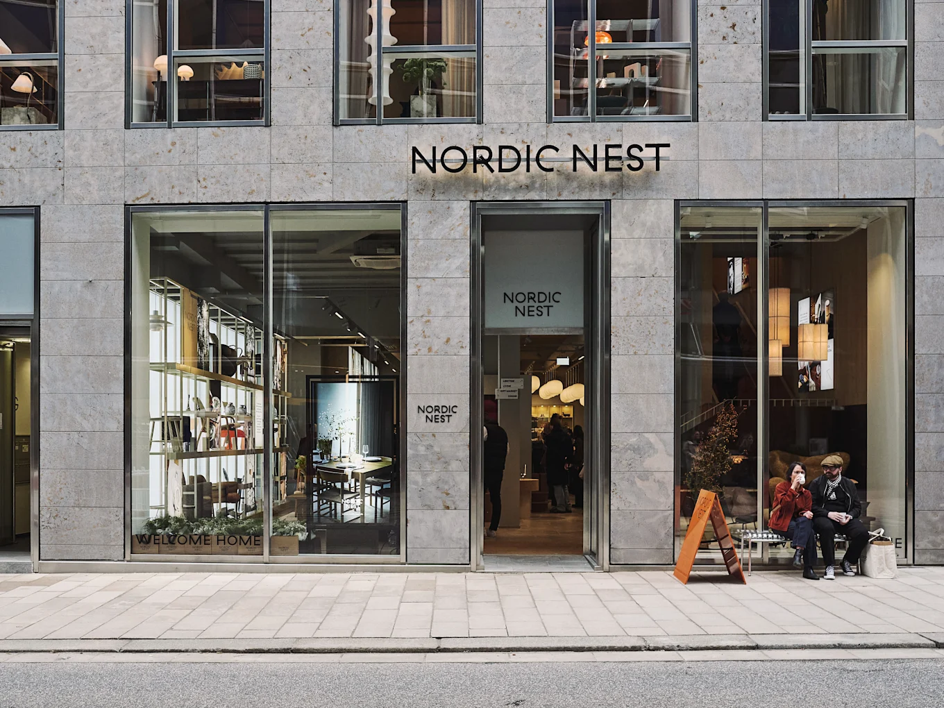 Moderne Nordic Nest butiksfacade med store vinduer, der udstiller boliginteriør, og to personer, der sidder på en bænk udenfor.