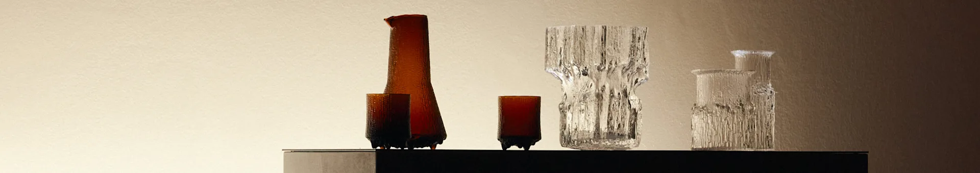 En samling teksturerede glasvaser og flasker, ravfarvede og klare, på en mørk overflade mod en lys baggrund.