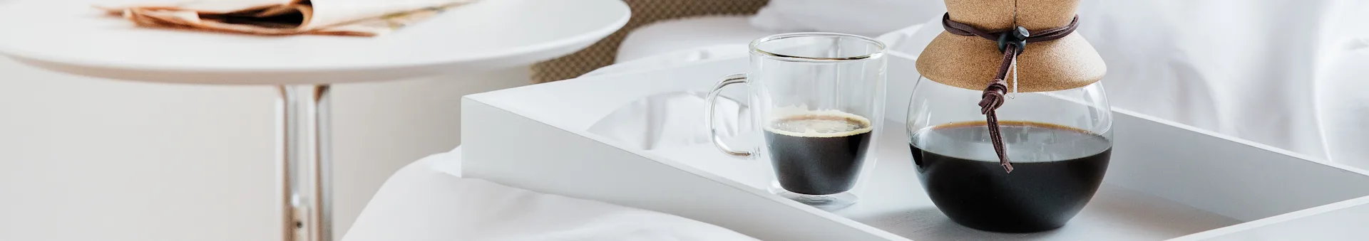En hvid bakke med et glaskrus kaffe og en kaffekande med en læderkrave på en seng.