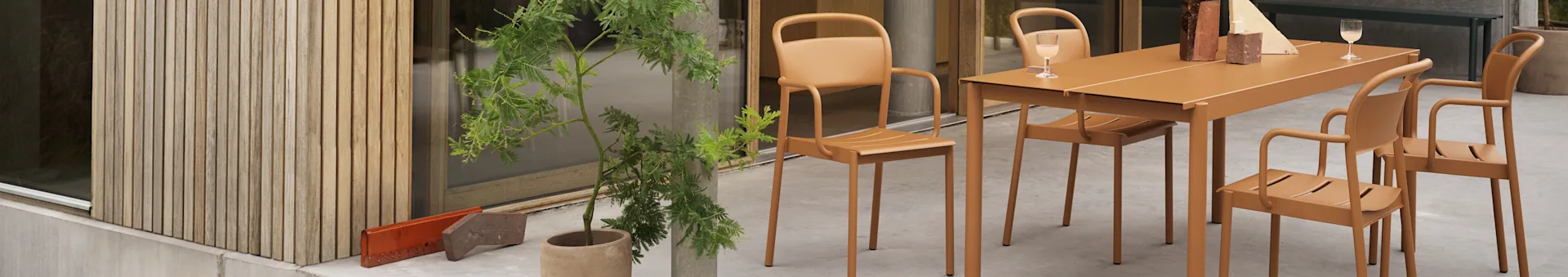 Orange udendørs spisebord med fire matchende stole på en terrasse ved siden af en træbeklædt væg og en grøn potteplante.