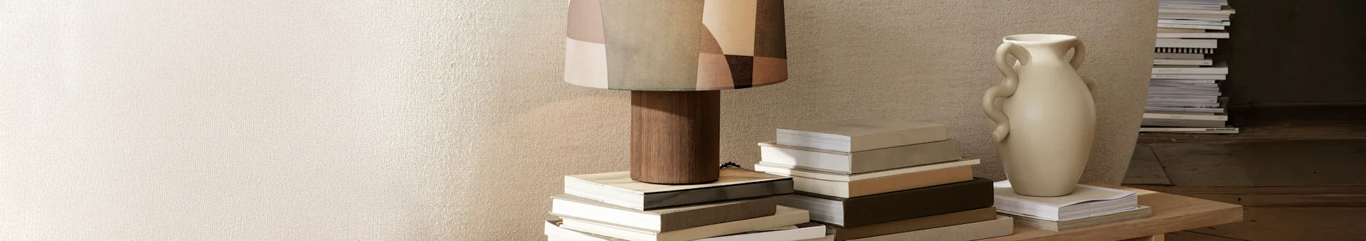 En høj lampe med en mønstret stofskærm og en lys beige vase står på et træbord, omgivet af stakke af bøger mod en tekstureret væg.