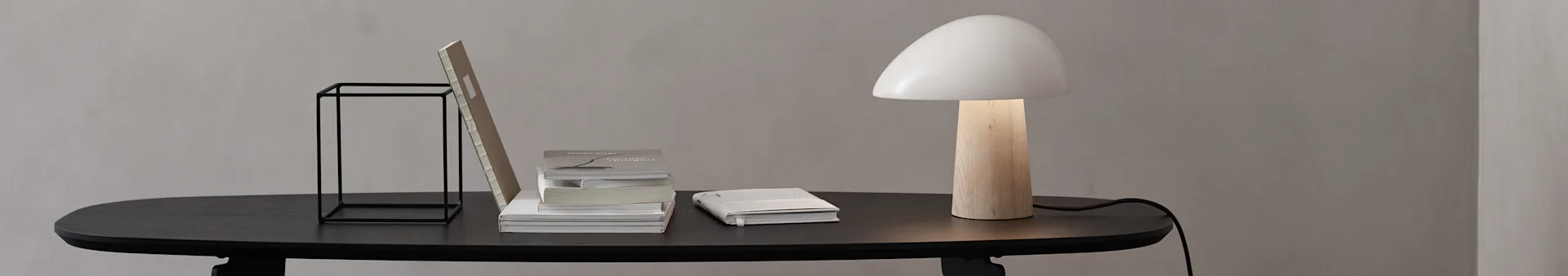 Et minimalistisk skrivebord med sort bord, sort metalstativ, stablede bøger og en lampe med træfod og hvid champagneskærm.