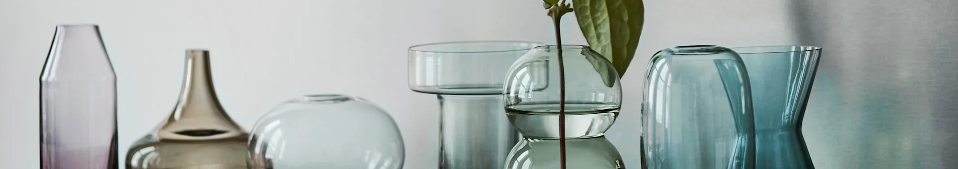 En række glasvaser og -flasker i lilla, brun, klar og blågrønne nuancer, hvoraf en holder en plantestilk.
