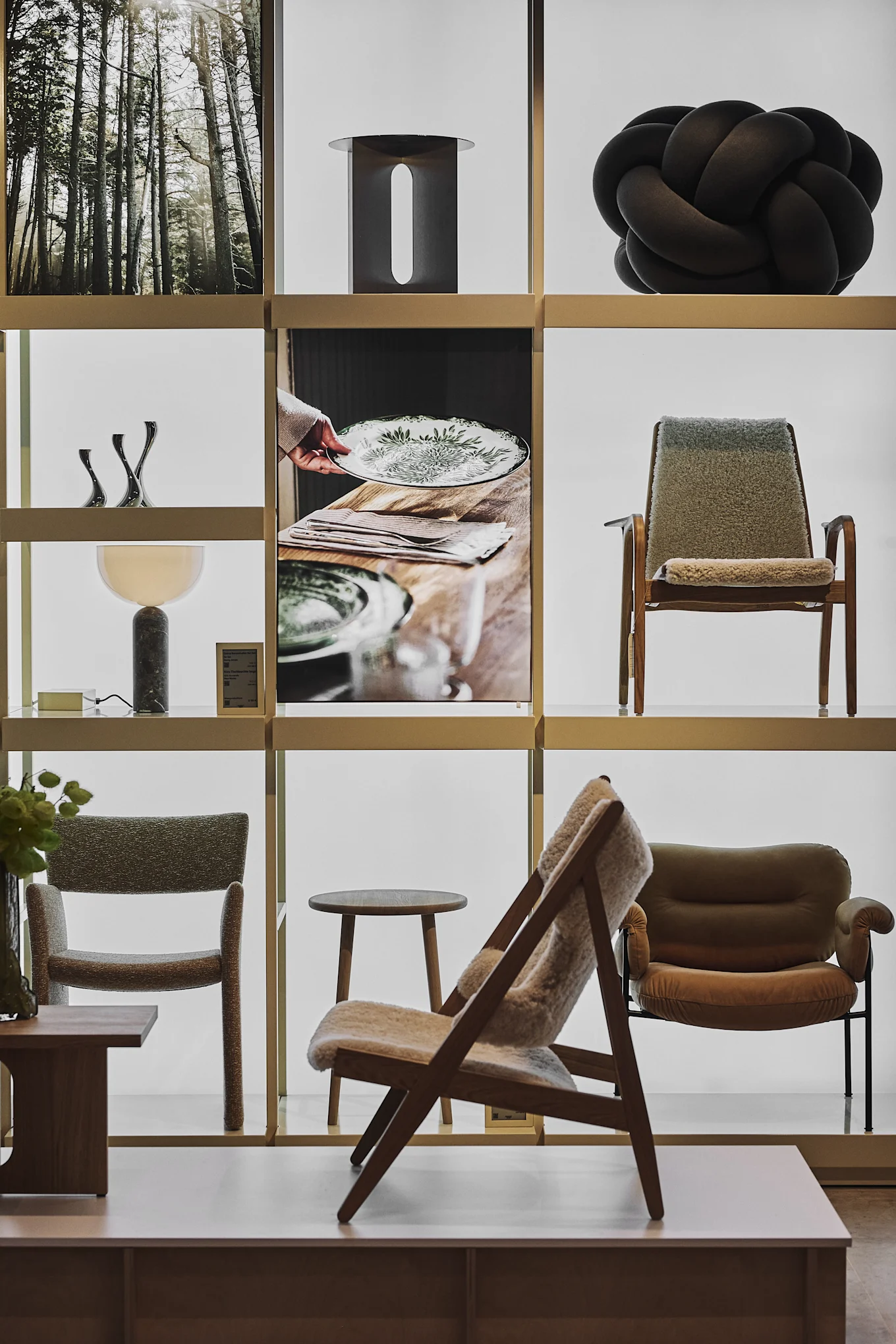 En hyldedisplay med boligindretning: et skovprint, sort knudeskulptur, metal kunst, en bordlampe og forskellige stole.