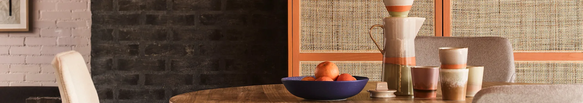 Et spisebord med en blå skål med citrusfrugter og gulerødder, omgivet af keramiske kander og krus, mod en flettet skærm og murstensvæg.