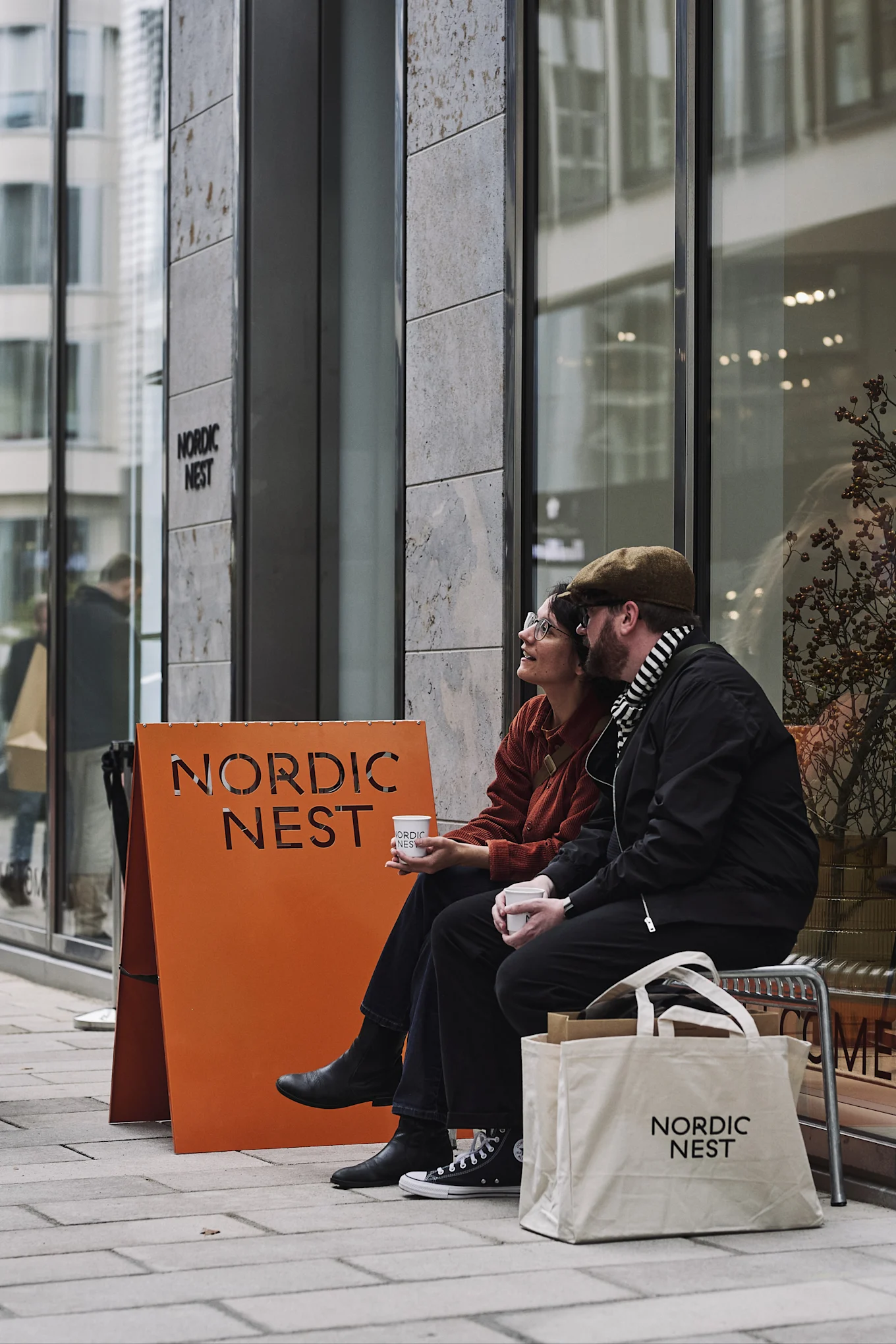 To personer sidder uden for en butik og holder kaffekopper med 'Nordic Nest'-logoet. Et orange skilt og en hvid mulepose viser også logoet.