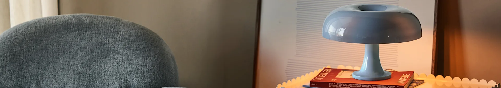En blå bordlampe står på et magasin ved siden af en gråpolstret stol.
