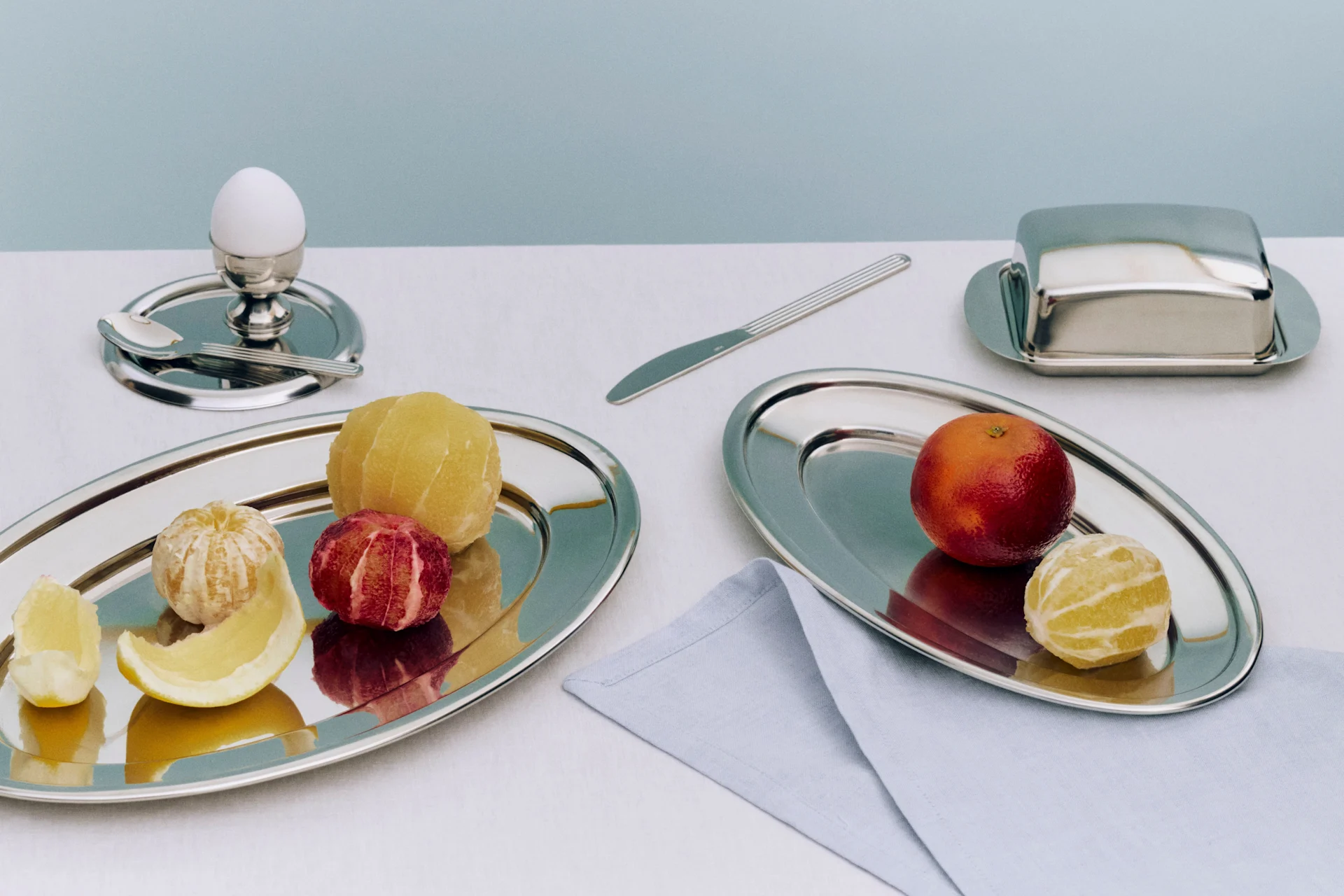 Morgenmad med pillede og upillede citrusfrugter på sølvbakker, et æg i en æggekop og en smørboks.