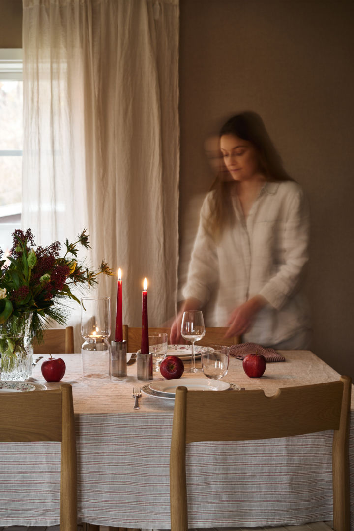 En kvinde dækker et enkelt julebord med røde stearinlys, mønstret juleporcelæn og en enkel stribet dug. 