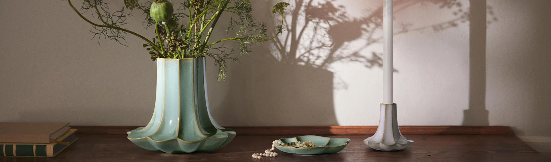 En riflet tealfarvet vase med løv, en perlekæde i en matchende skål, et hvidt lys i en riflet holder og bøger på en træoverflade.