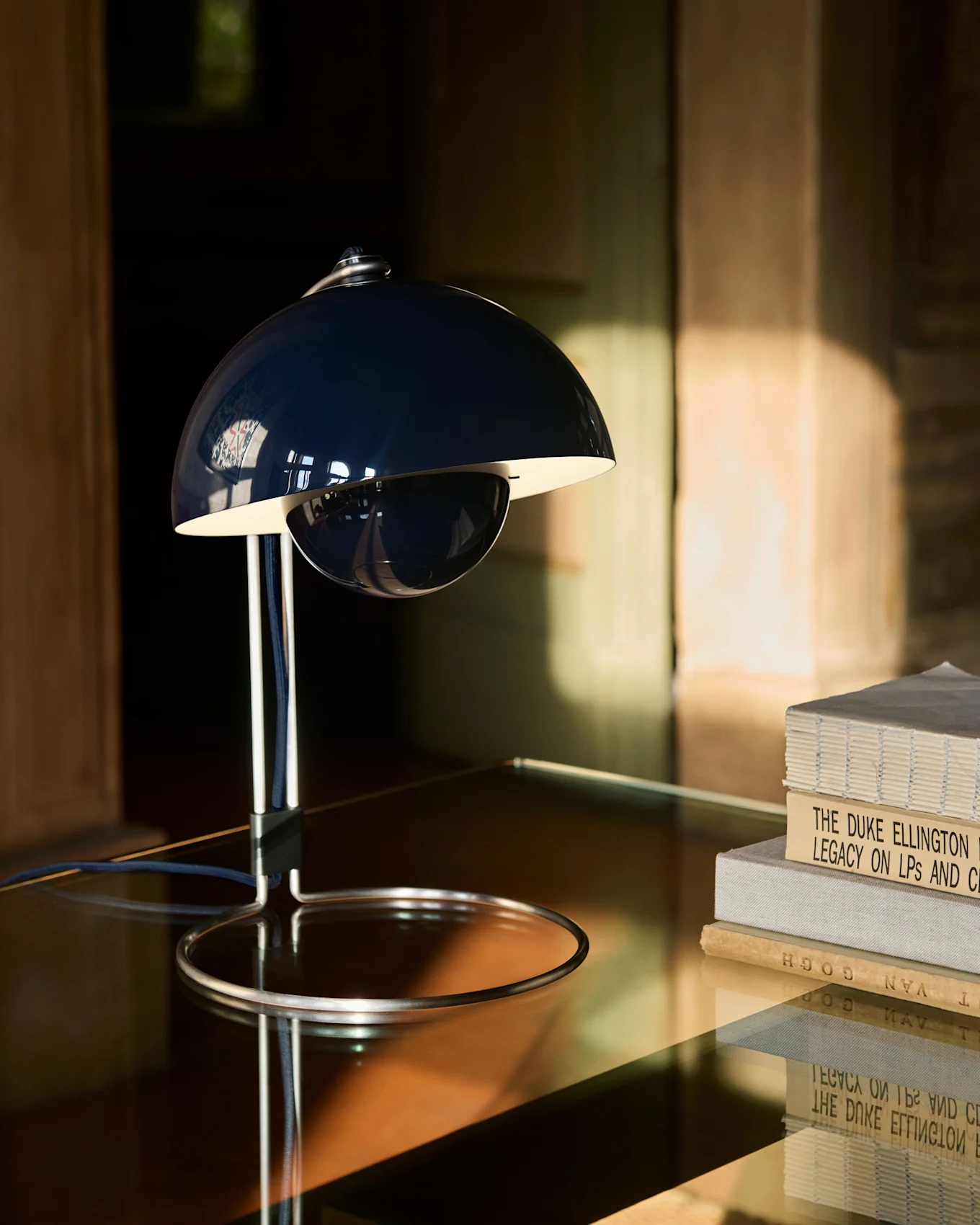 En mørkeblå, klokkeformet bordlampe med en kromfod står på et reflekterende bord ved siden af en stak bøger.