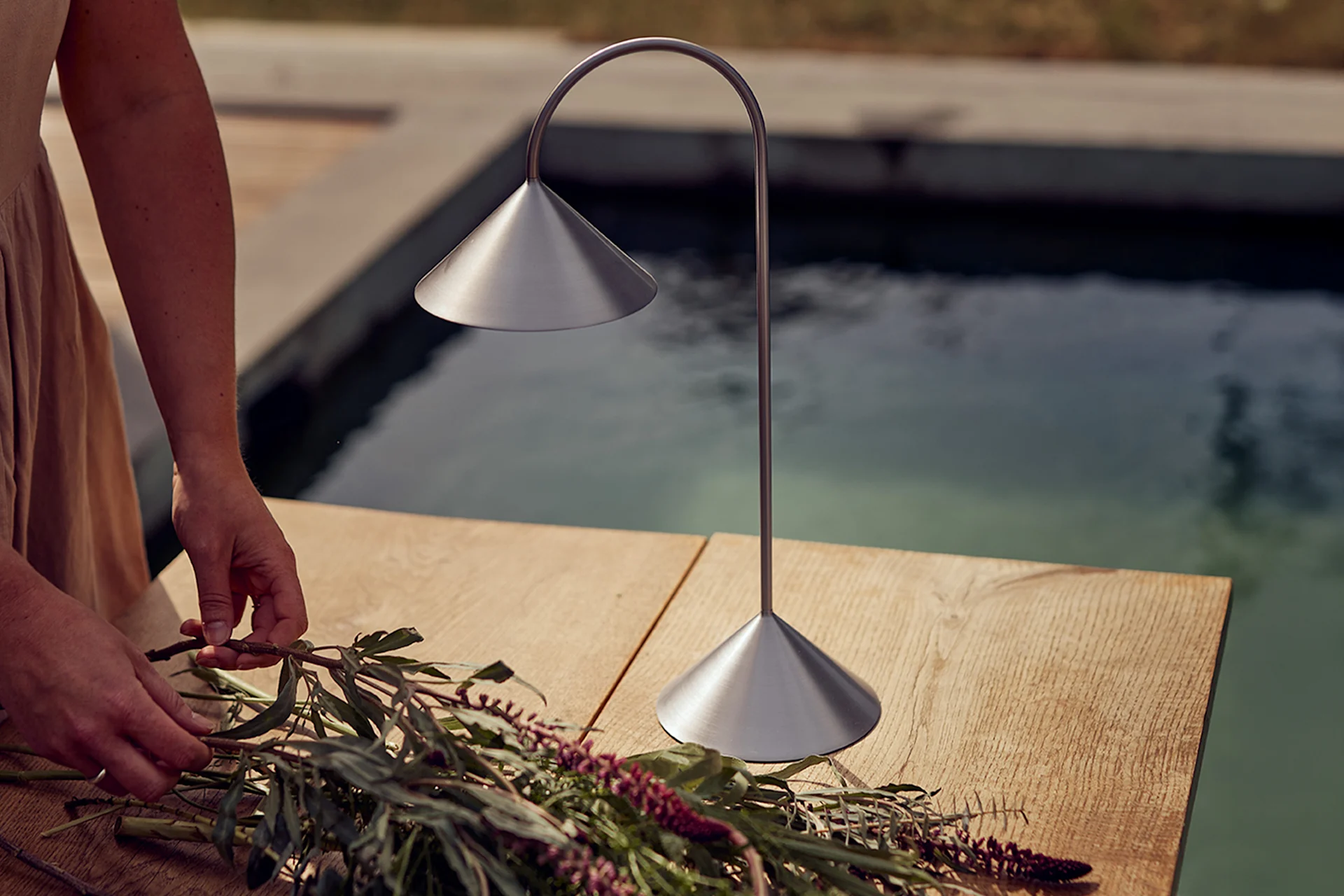Hænder arrangerer blomster på et træbord ved siden af en børstet metalbordlampe ved en pool.