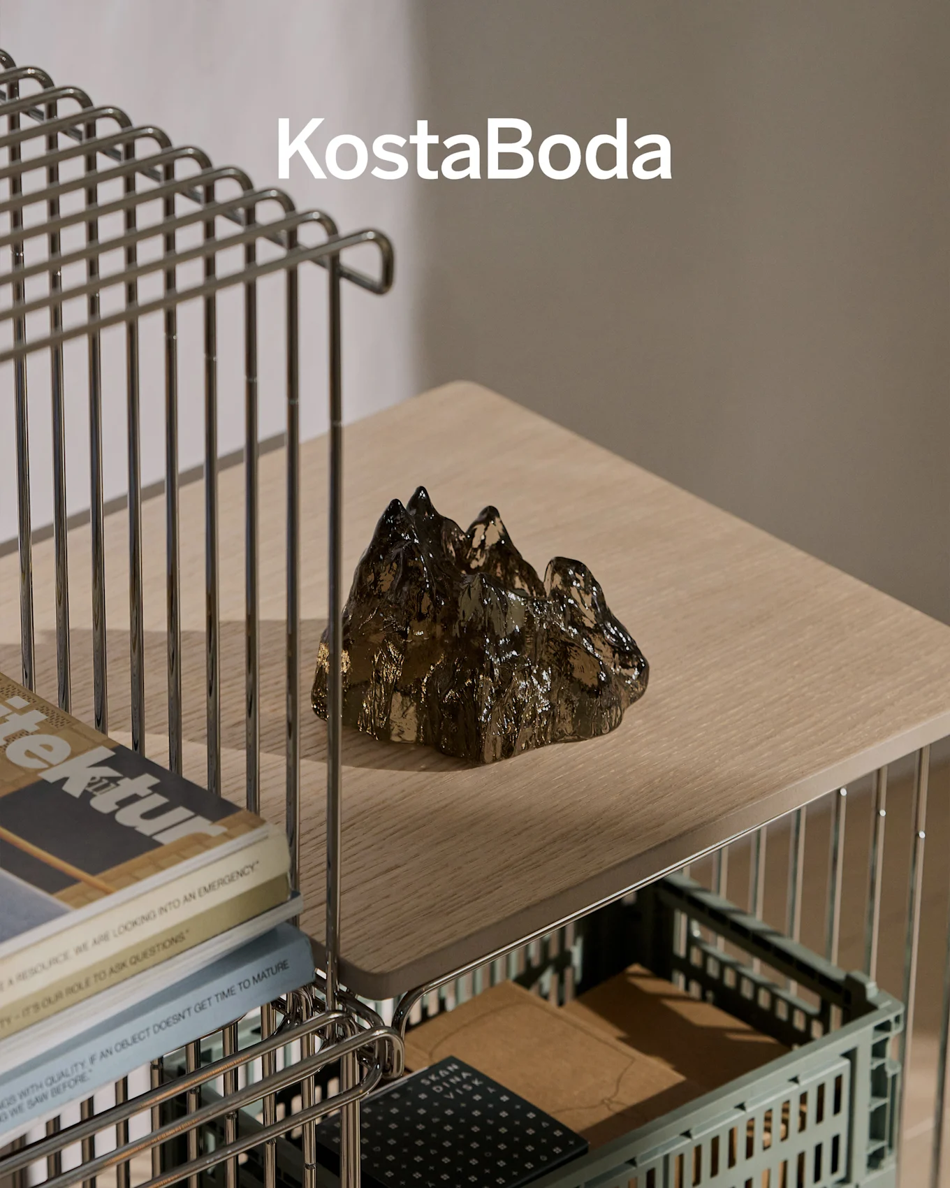 KostaBoda brandnavn over en mørk, transparent, bjergformet glasskulptur på en lys træhylde.