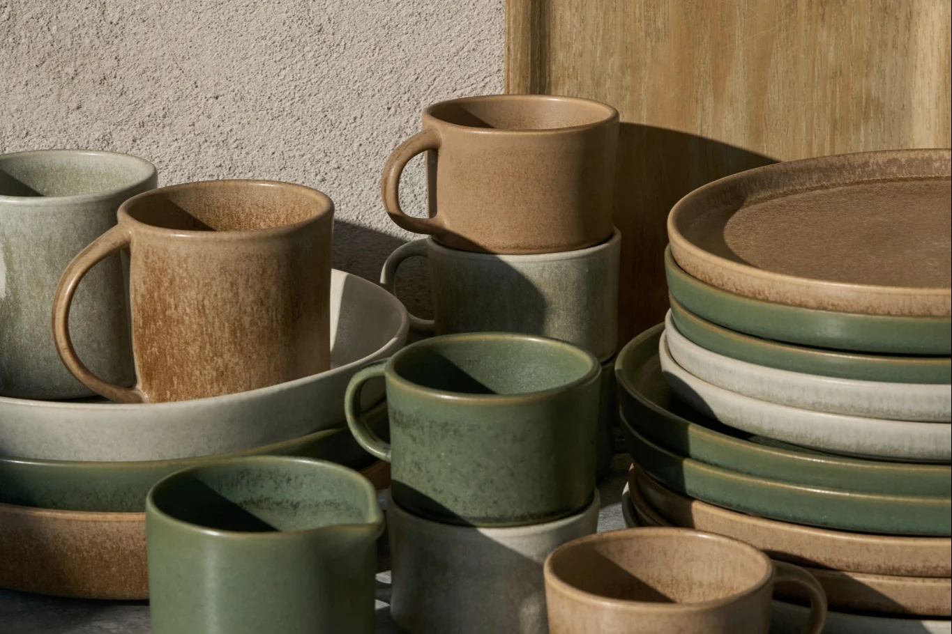 Stablede keramikkrus, skåle og tallerkener i beige, lysegrå og salviegrøn med en rustik finish.