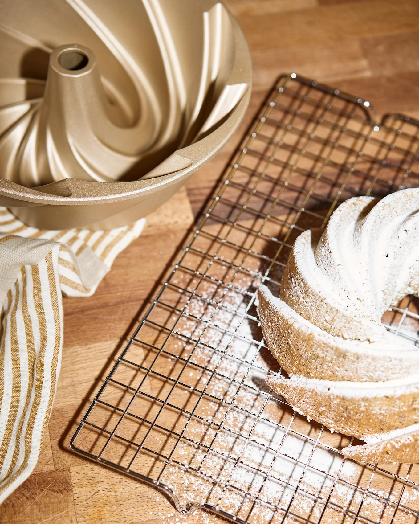 En gylden bundt kageform, en kølerist med en flormelis-dækket bundtkage og et stribet håndklæde på en træoverflade.