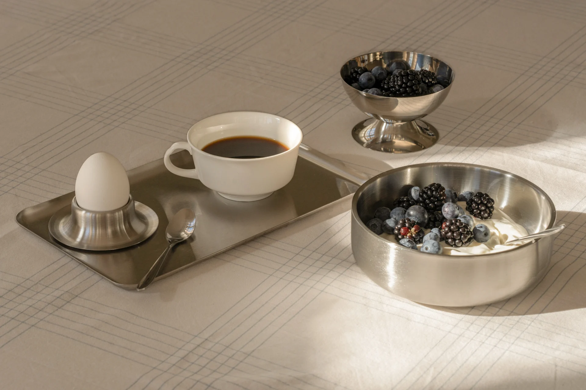 Morgenmad med kaffe, kogt æg, yoghurt og friske bær i sølvskåle på en mønstret dug.
