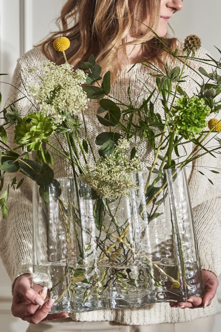 En kvinde i creme står og holder Gry wide vasen fyldt med vilde forårsblomster.