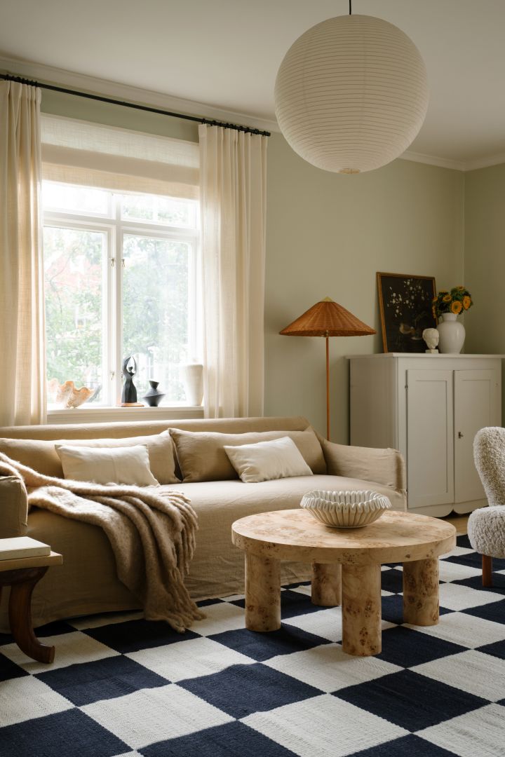 Et billede der viser Rice papir lampeskærmen fra HAY, den hænger i en stue med en beige sofa, sort og hvidt ternet gulvtæppe og et rustiks kaffebord.