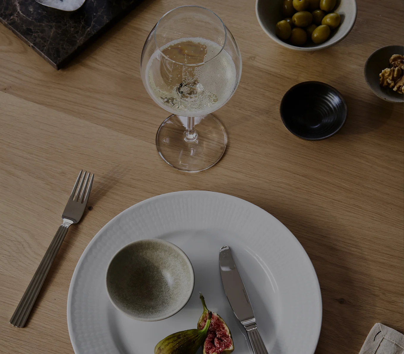 Et glas champagne, hvid tallerken med skål og figner, gaffel, kniv, oliven og valnødder på et træbord.