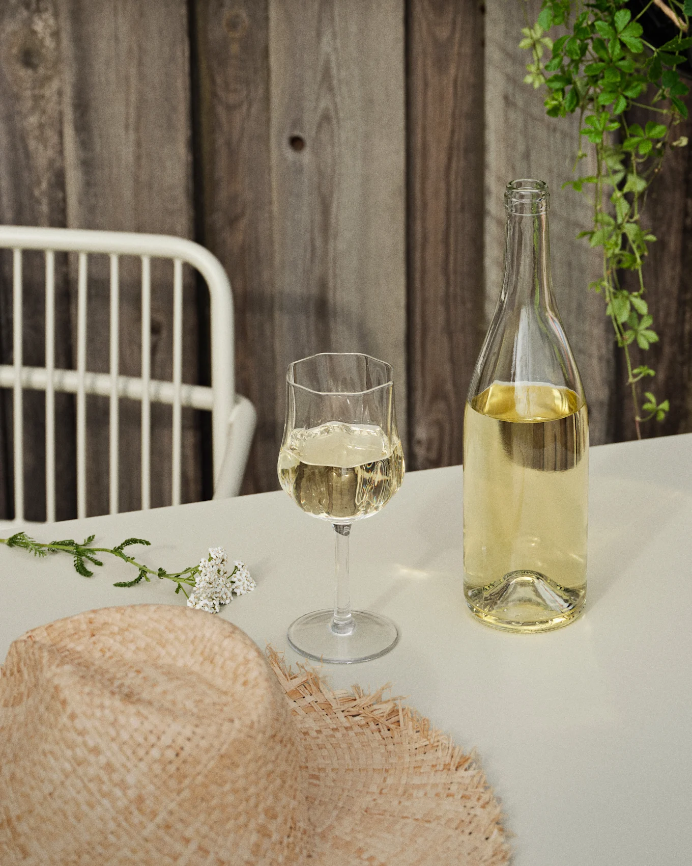 En flaske og et glas hvidvin på et bord med en stråhat og hvide blomster, mod en trævæg og grønne slyngplanter.