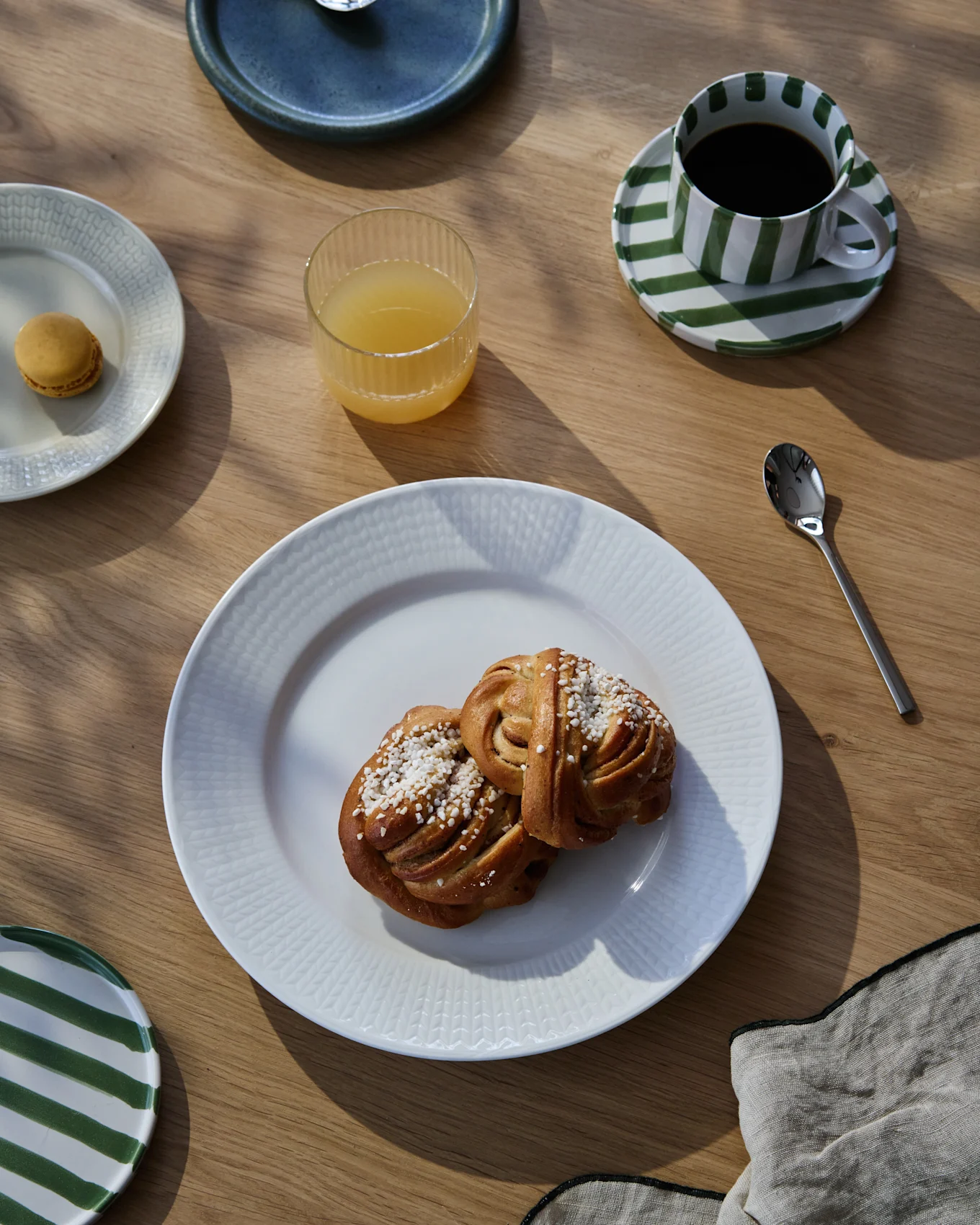 En morgenmadsanretning på et træbord med to kanelsnegle, kaffe i en stribet kop, appelsinjuice og en makron.