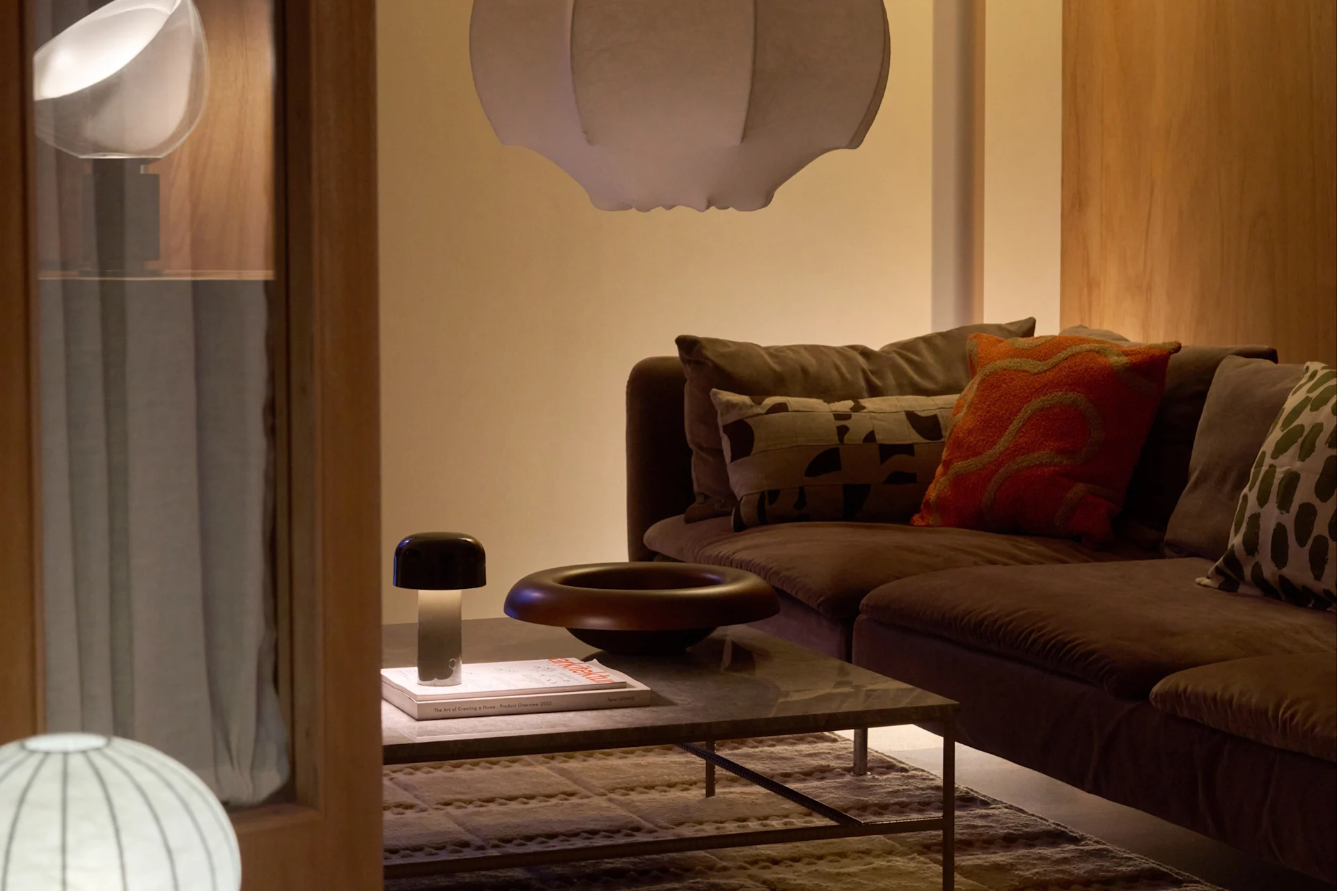 En hyggelig stue med brun sofa, hvid pendellampe, gulvlampe og sofabord med champignonlampe og skål.