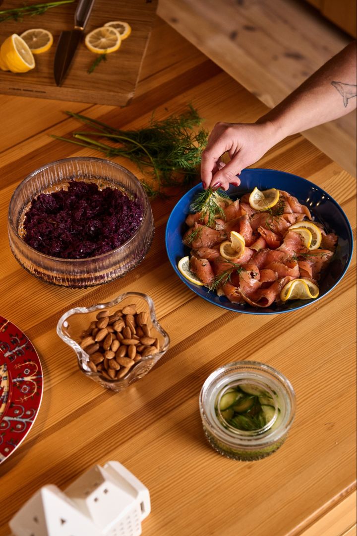 En hånd lægger et stykke dild på en blå tallerken med laks og citron, en traditionel ret som en del af den nordiske julefejring.