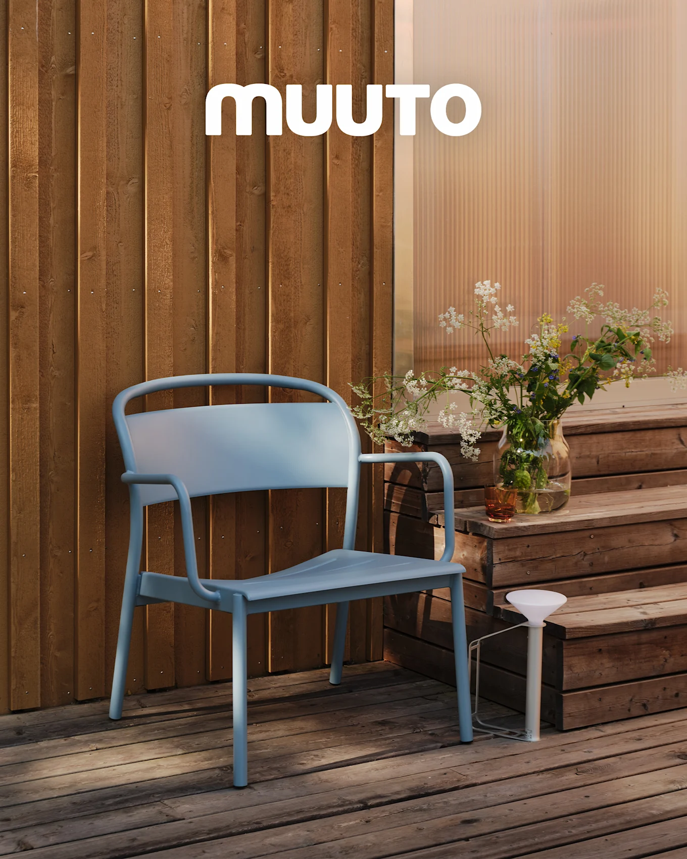 En lyseblå udendørsstol på et trædæk ved trin med en vase vilde blomster og en hvid bærbar lampe. MUUTO logo.