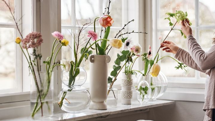 En samling af vaser i klart glas med farverige forårsblomster der står i vindueskarmen.