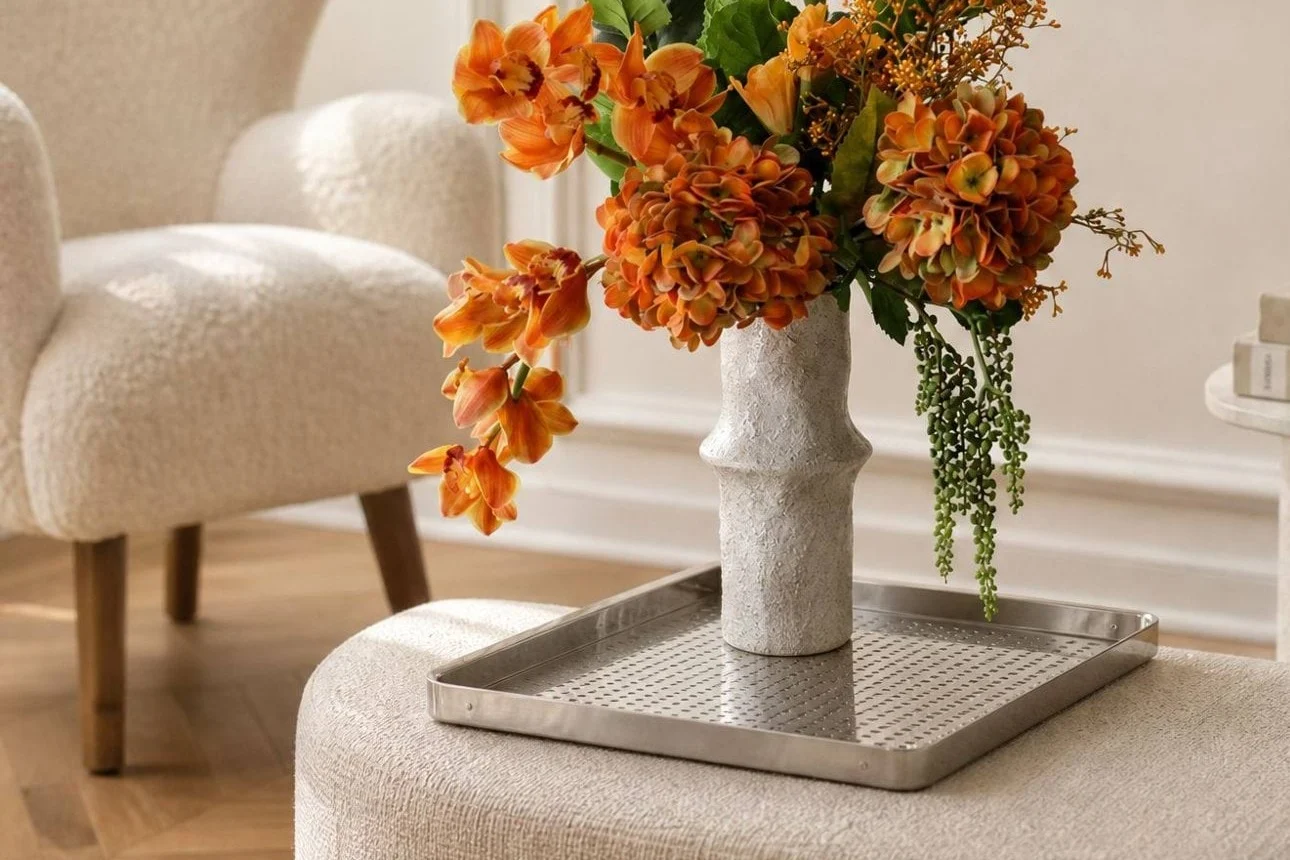 Levende orange blomster i en tekstureret hvid vase på en sølvbakke, placeret på en lysegrå puf i en moderne stue.
