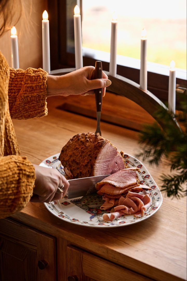 En kvinde skærer en traditionel nordisk juleskinke i et vindue ved siden af en julelysbue.