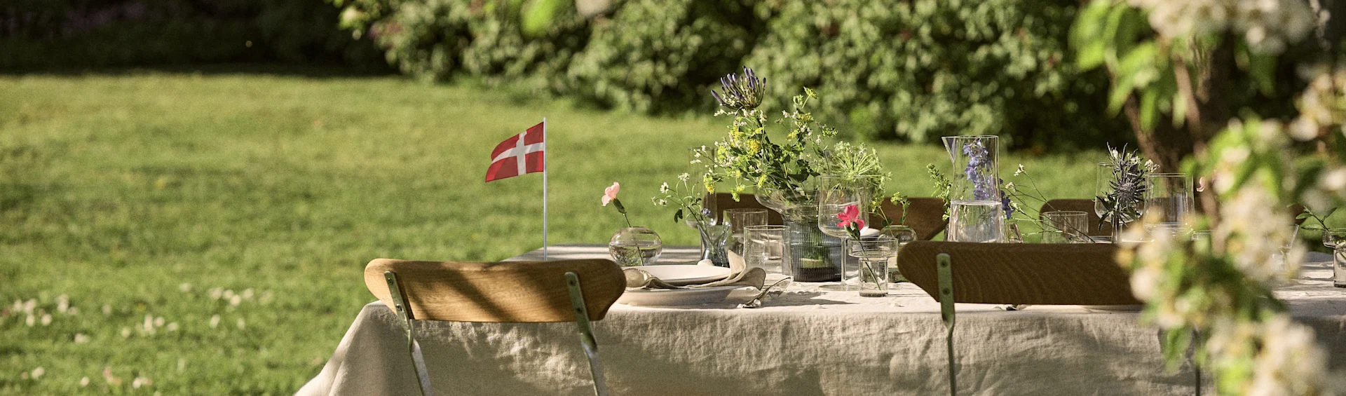 Et dækket bord udendørs med blomster, glas, tallerkner og et lille dansk flag i en grøn have.