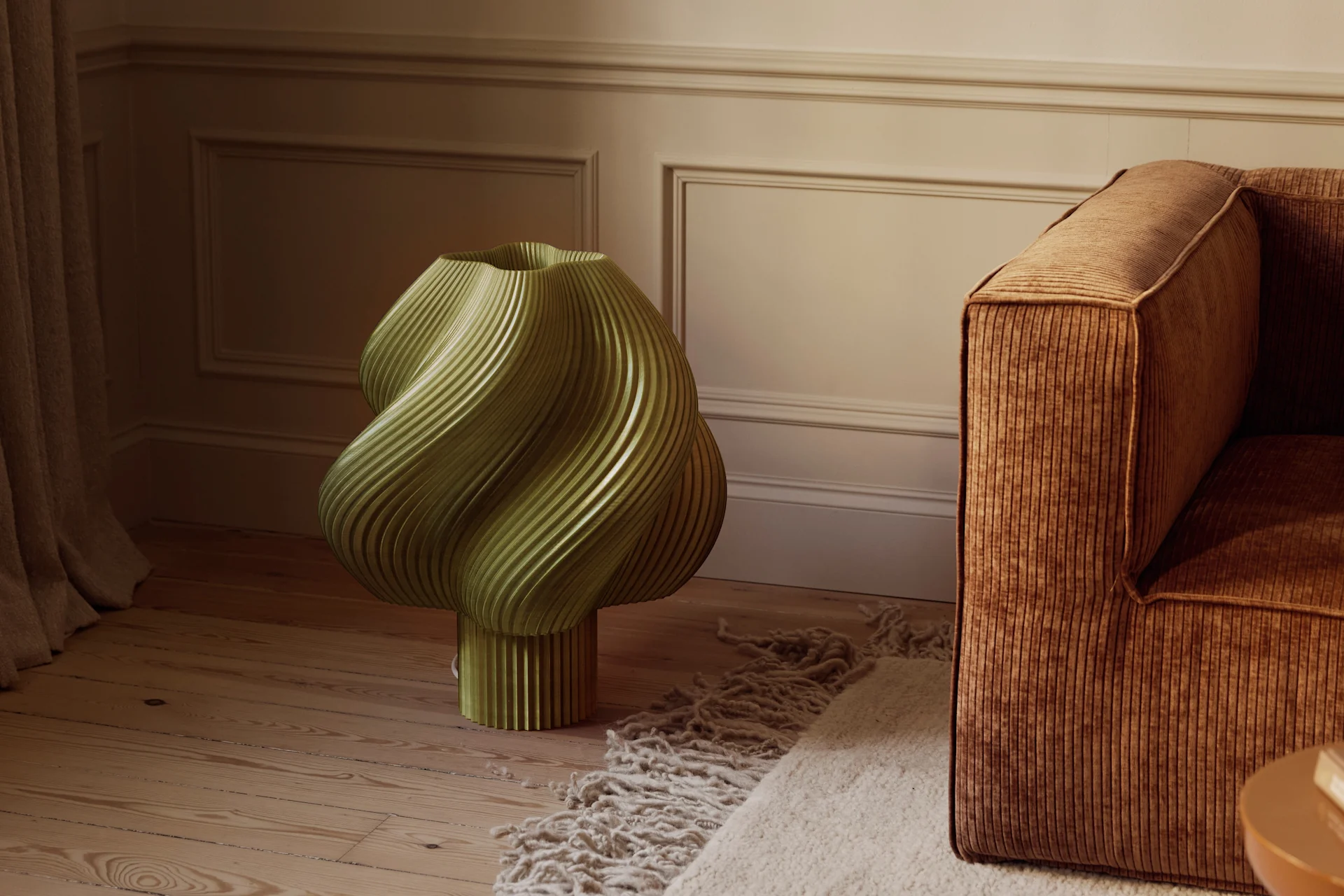 En olivengrøn, skulpturel, riflet genstand på et trægulv ved siden af en brun fløjlssofa og et tekstureret tæppe i et rum med profilerede vægge.